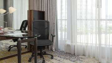 A hotel room with a desk, office chair, lamp, and a large window with sheer curtains.