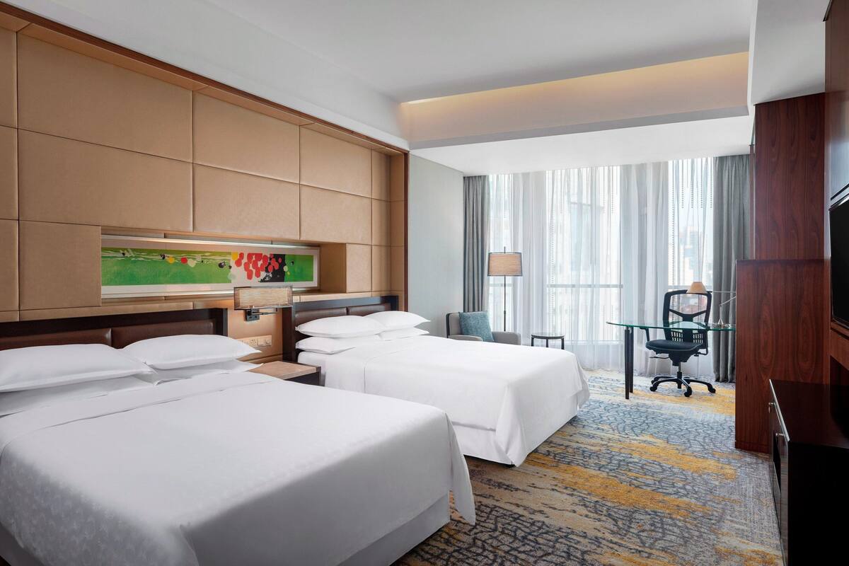 A hotel room with two beds, a desk with a chair, a television, and a large window with curtains.