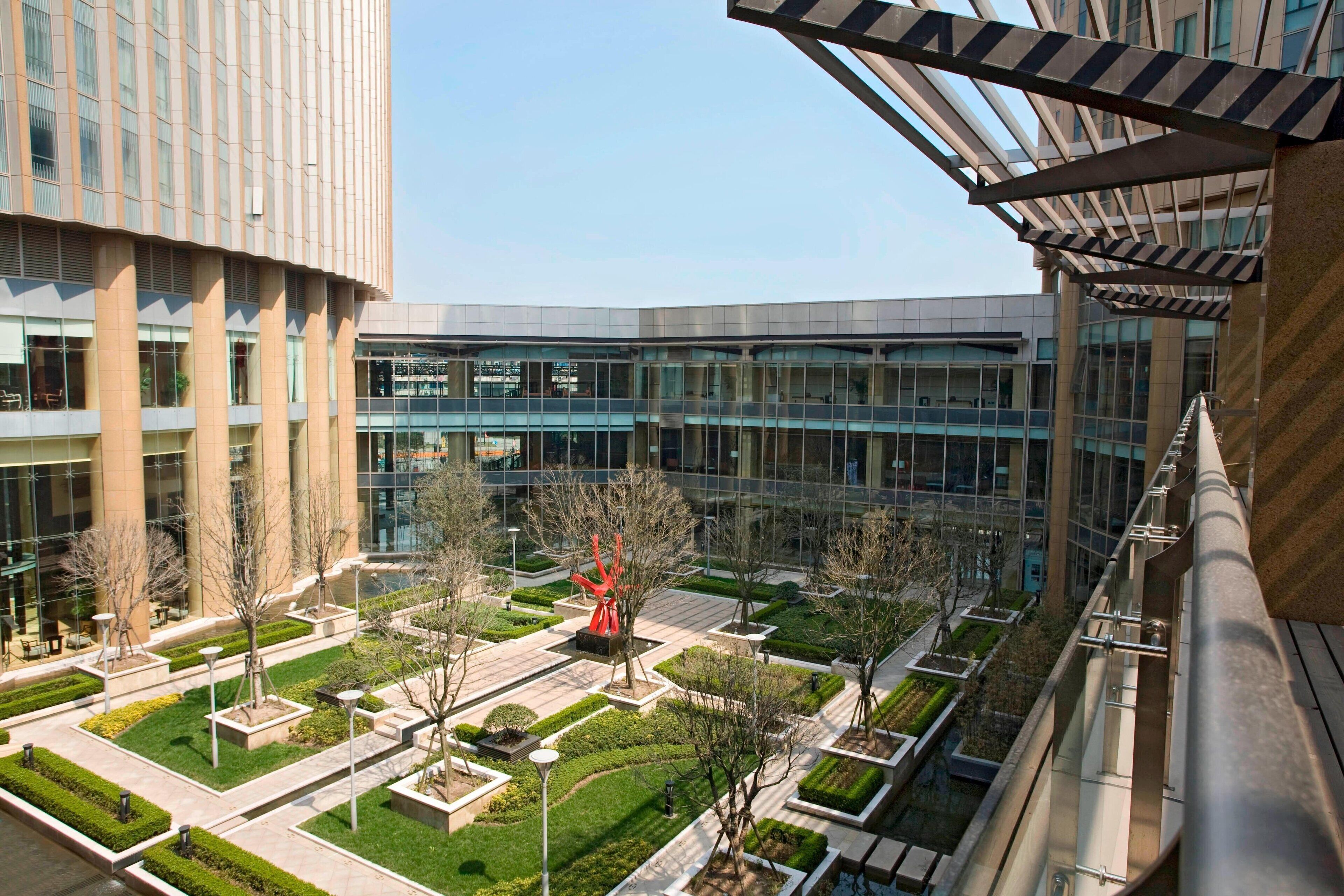A modern building with a courtyard featuring geometric landscaping, a prominent red sculpture, and a glass facade.