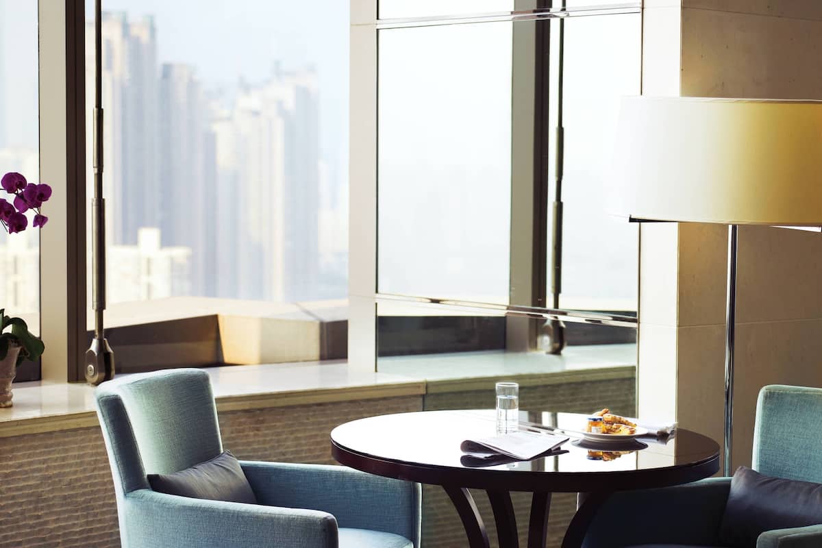 A modern hotel room with a large window offering a city view, a round table with a glass of water and a plate of food, and two blue upholstered chairs.