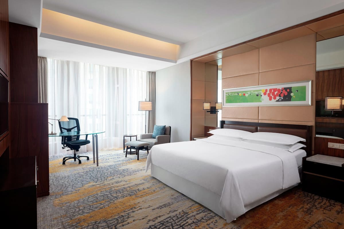 A hotel room with a large bed, a desk with a chair, a small table, and a large window with sheer curtains.