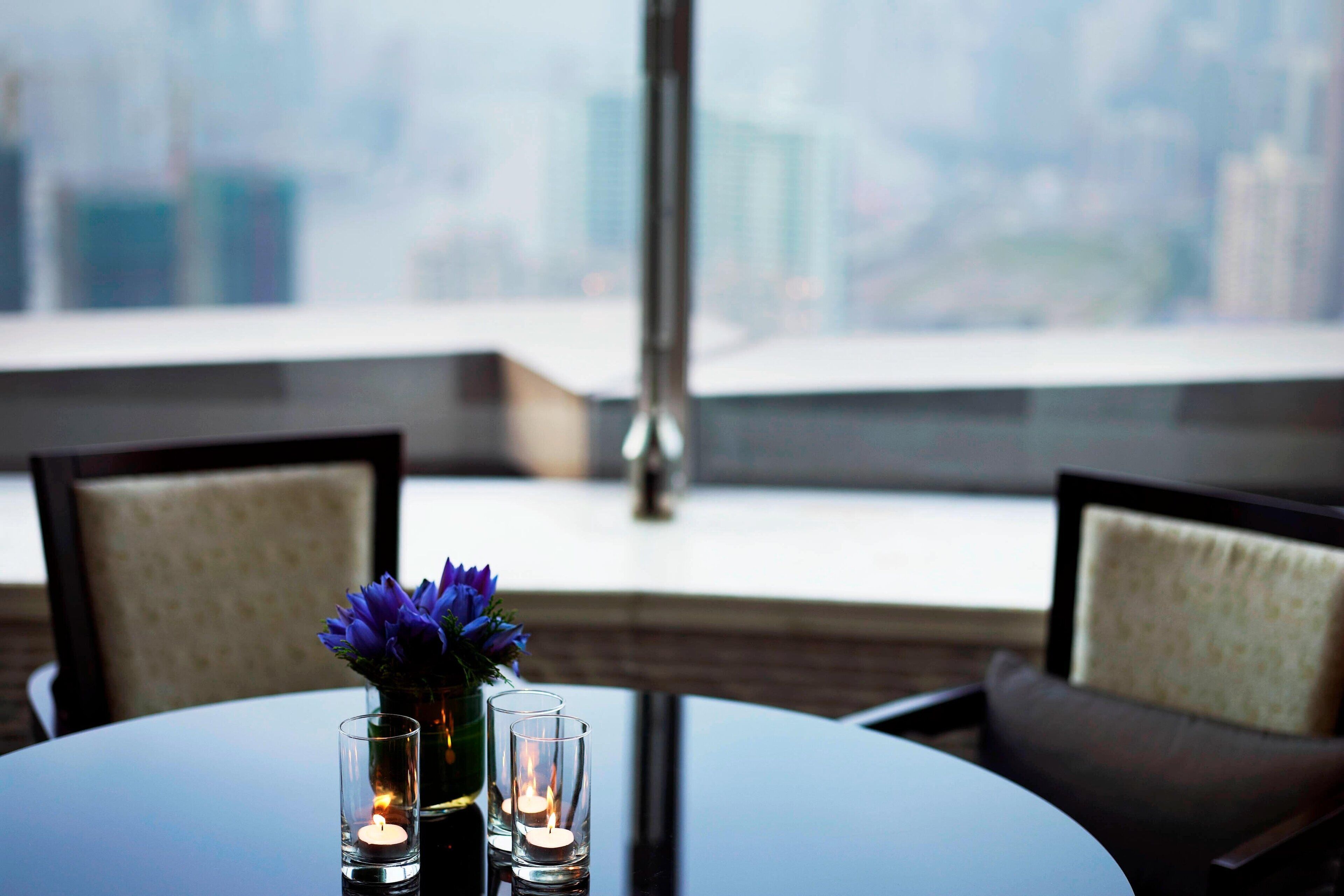 A round table with candles and a vase of flowers, set against a cityscape view.