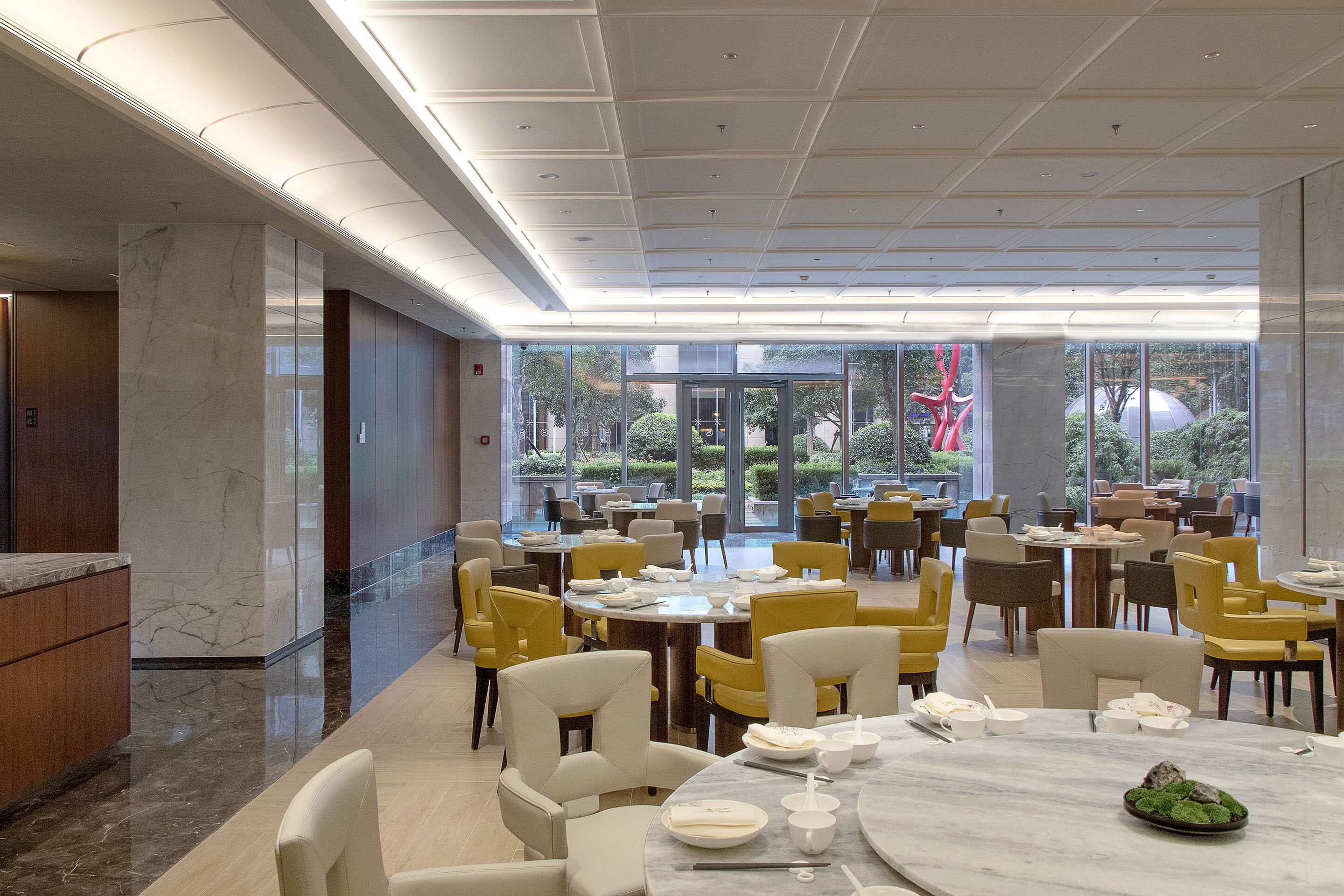 A spacious dining area with multiple tables and chairs, large windows offering views of greenery, and a modern ceiling design.
