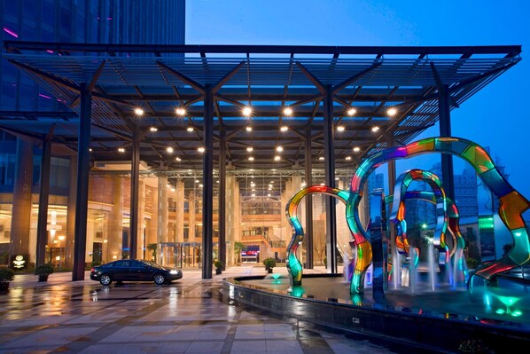 A modern hotel entrance with a glass facade, a covered outdoor area with a metal roof, and a colorful abstract sculpture in the foreground.