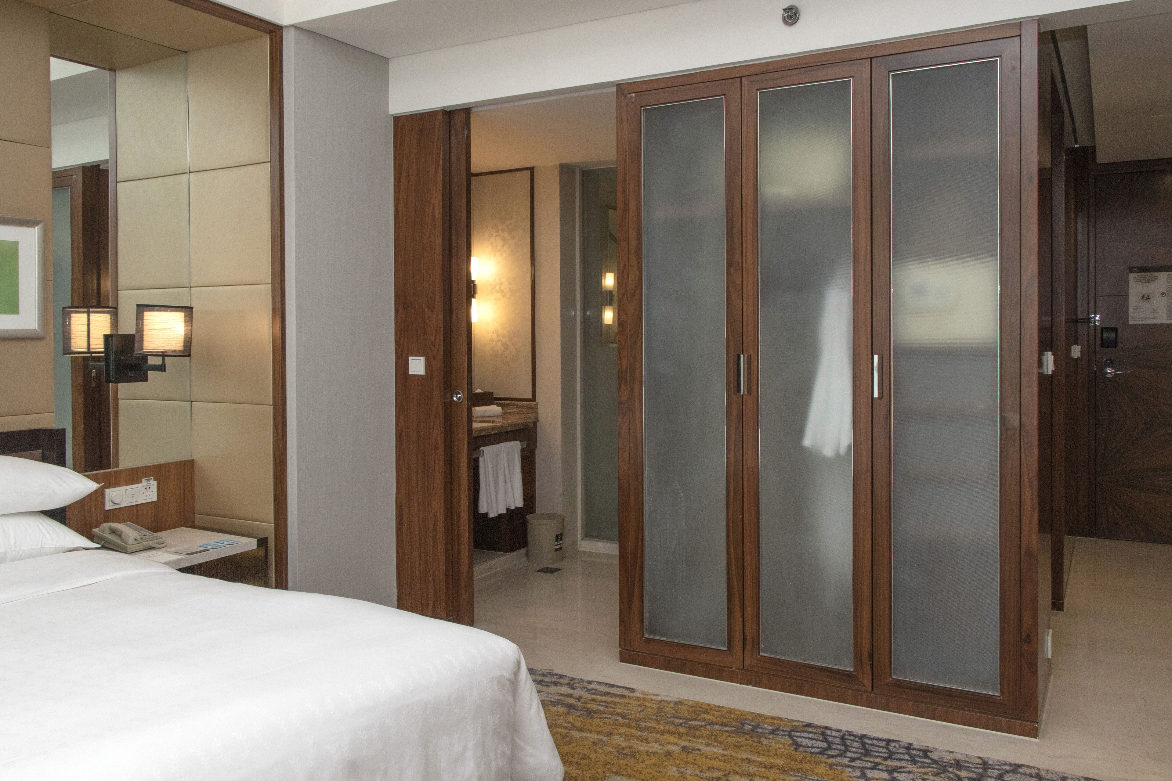 A hotel room with a bed, a bathroom with a mirror, and a wardrobe with frosted glass doors.