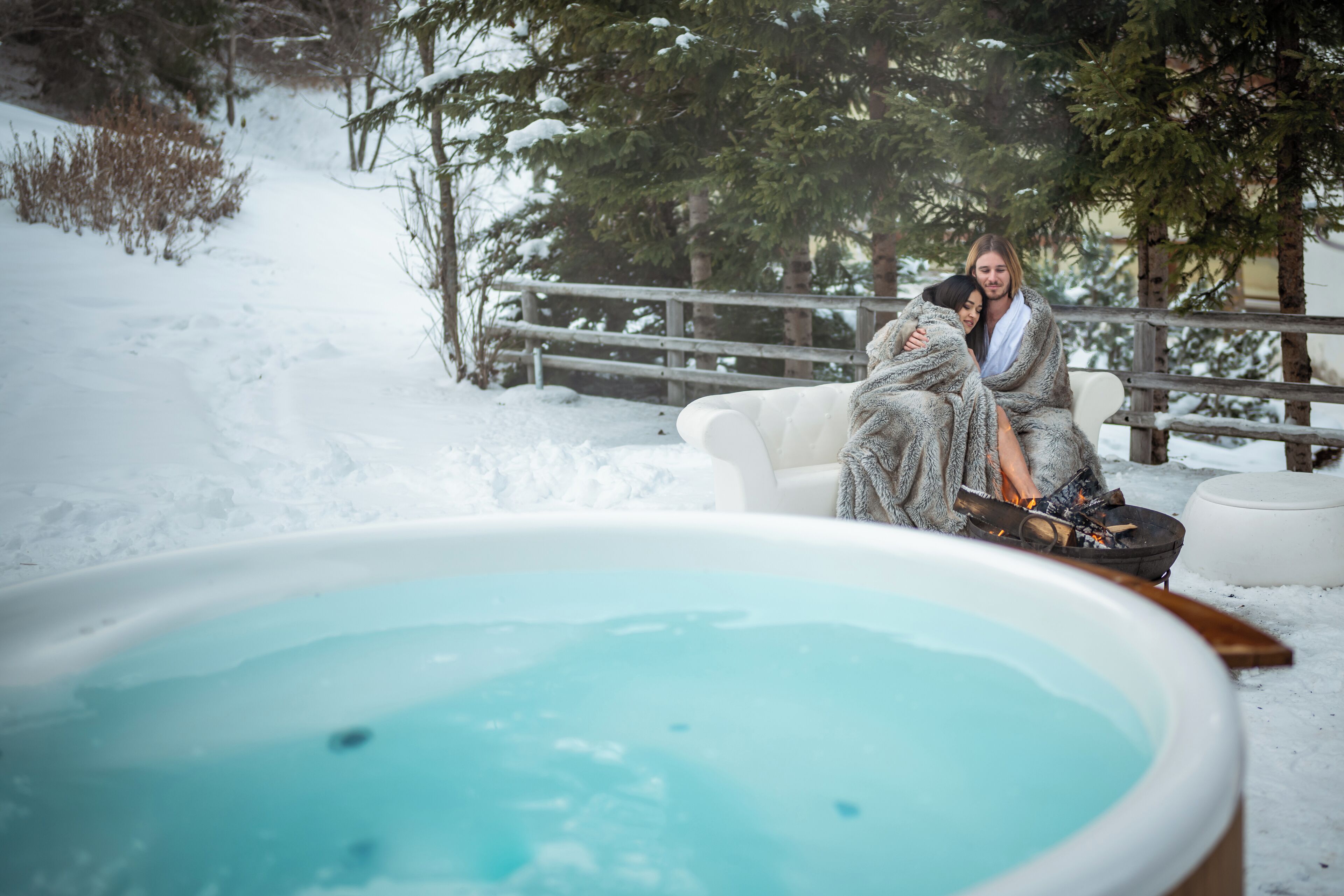 Un espacio exterior cubierto de nieve con un jacuzzi, bancos de madera y un pozo de fuego.