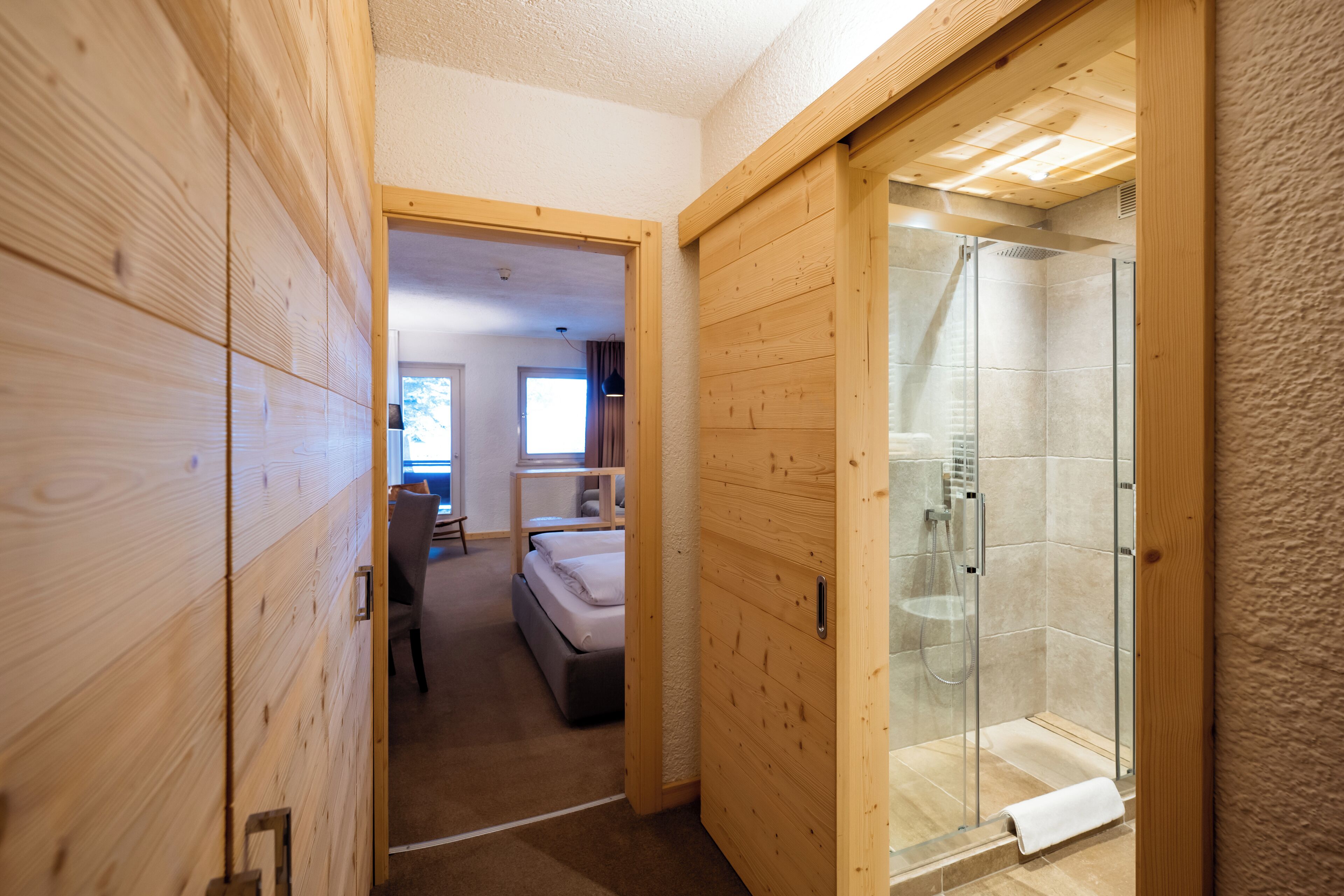 Un couloir menant à une chambre à coucher équipée d’un lit, d’une douche en verre et d’une porte en bois.