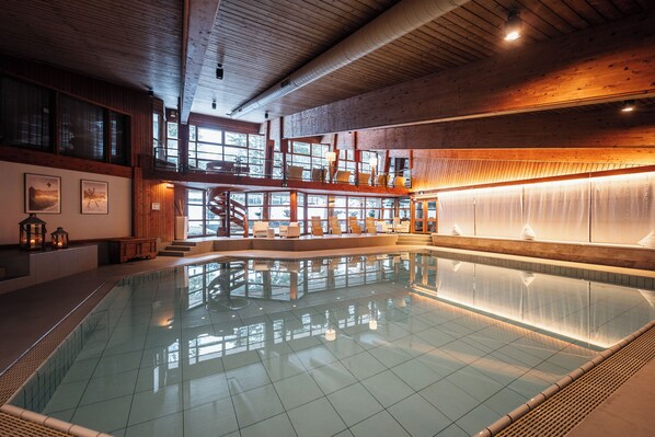 Una amplia piscina interior con techo alto y vigas de madera, rodeada de zonas de estar y una escalera que conduce a un nivel superior.