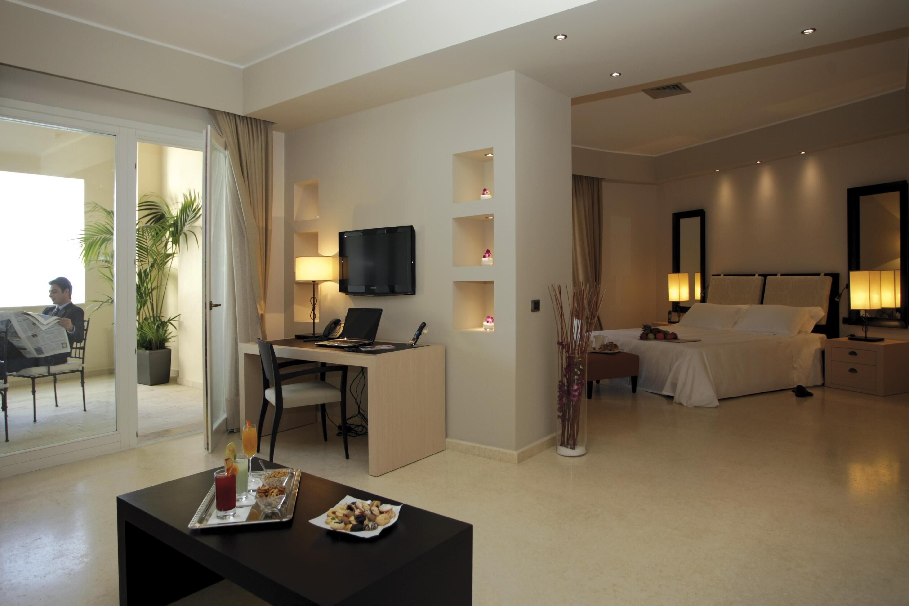 Una habitación de hotel moderna con escritorio, computadora, televisor de pantalla plana, cama, lámparas de noche y un área de comedor con una bandeja de comida y bebidas.