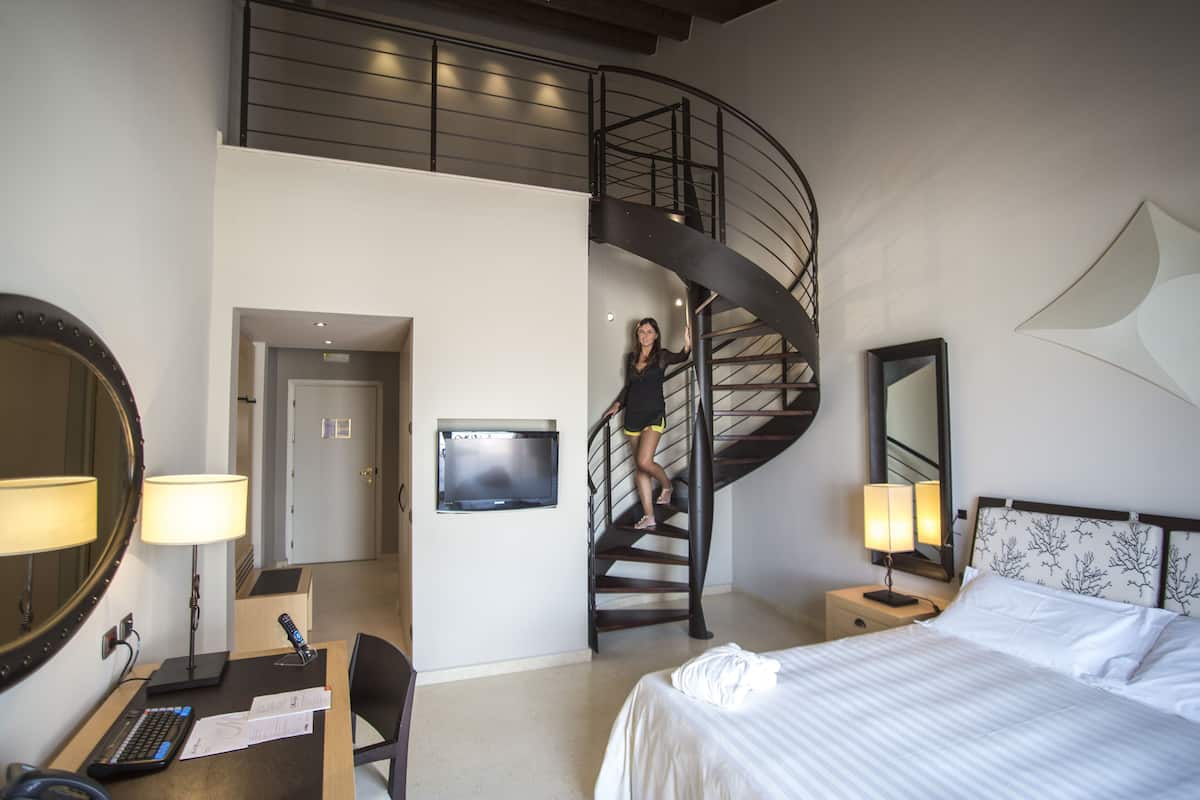 A hotel room with a bed, desk, chair, desk lamp, TV, and a spiral staircase.