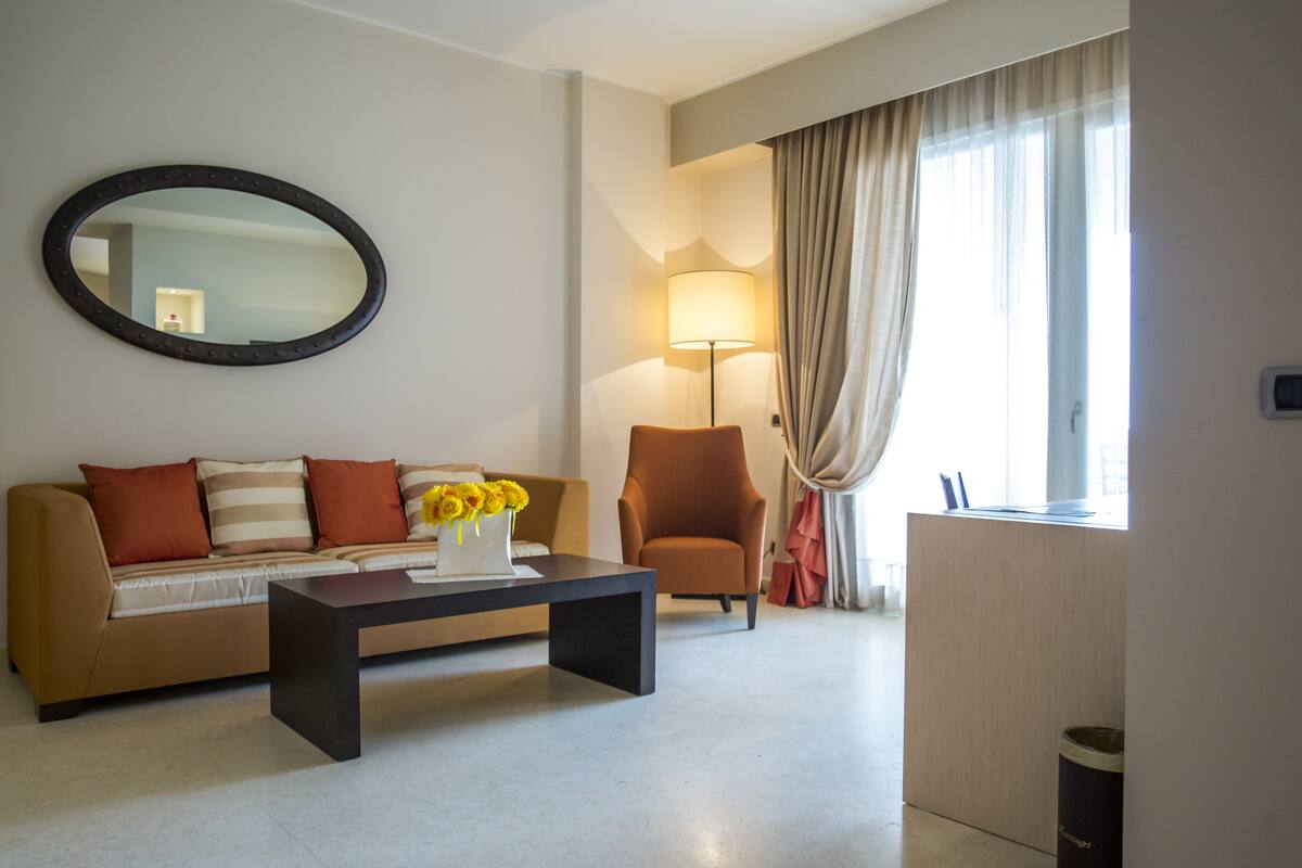 A modern hotel room with a sofa, armchair, and a coffee table with a vase of flowers.