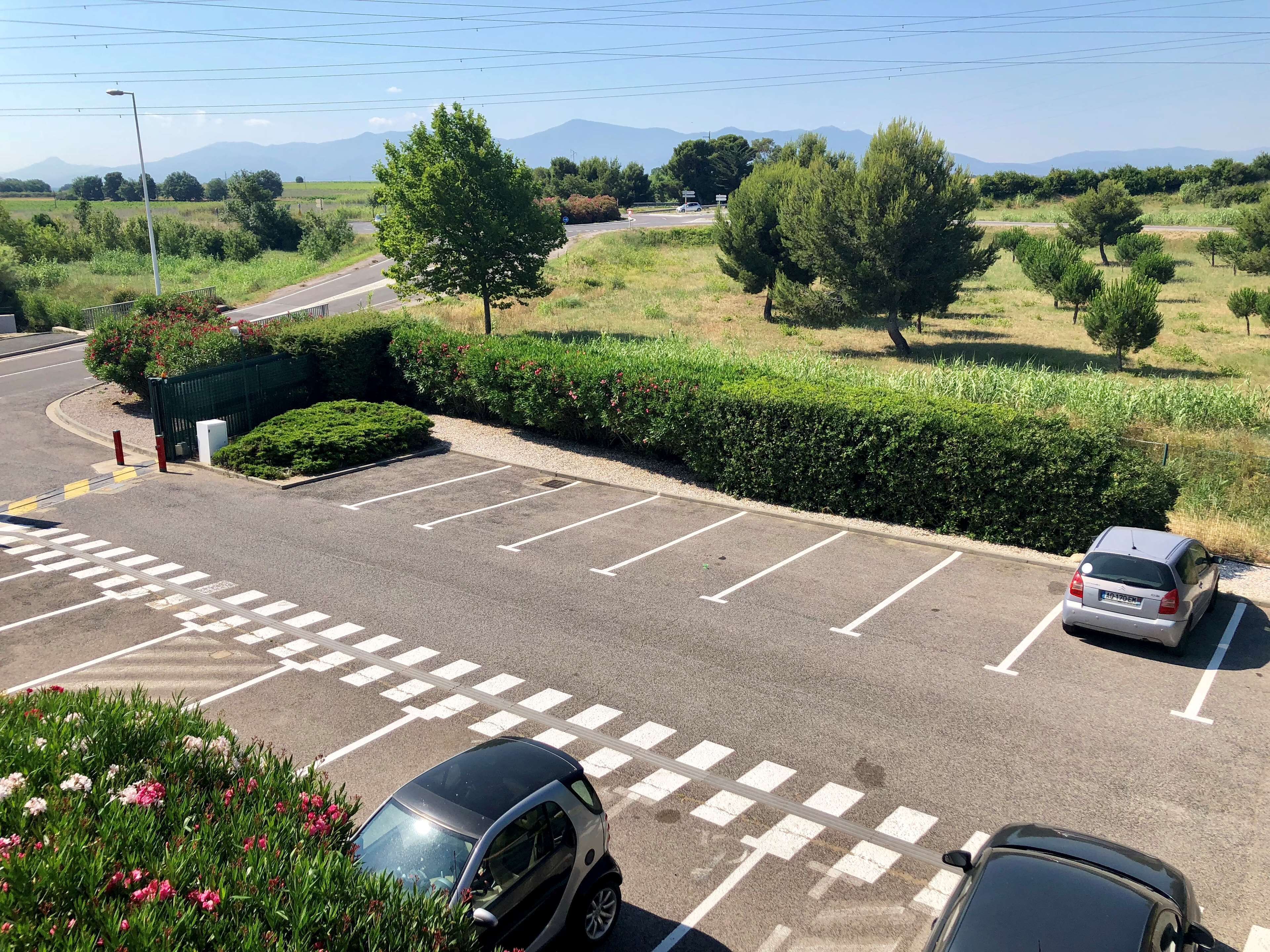 Um estacionamento com vagas demarcadas, uma faixa de pedestres e vista para uma estrada com veículos.