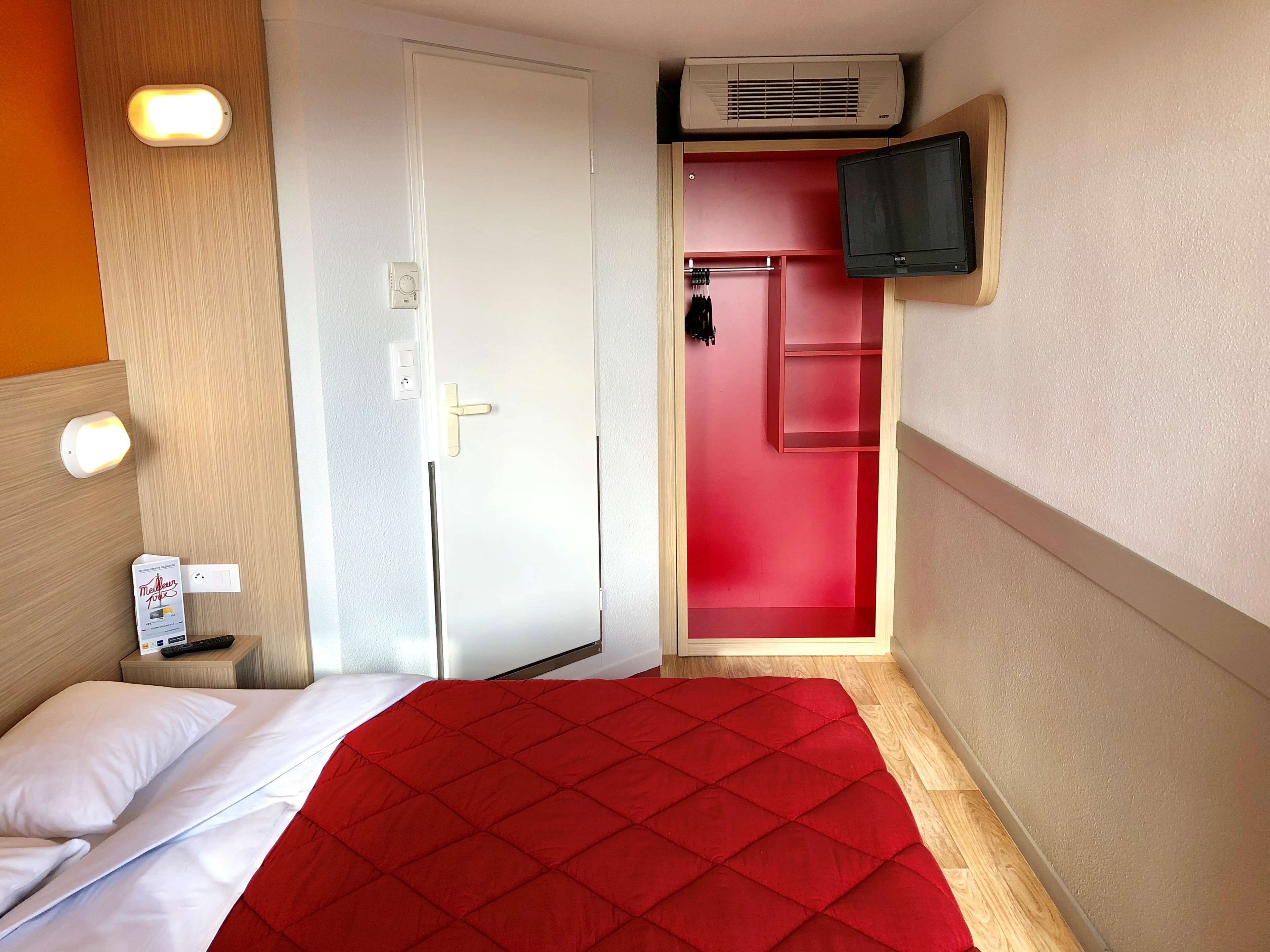 A hotel room with a bed, a red door, a mounted TV, and a wall-mounted air conditioner.
