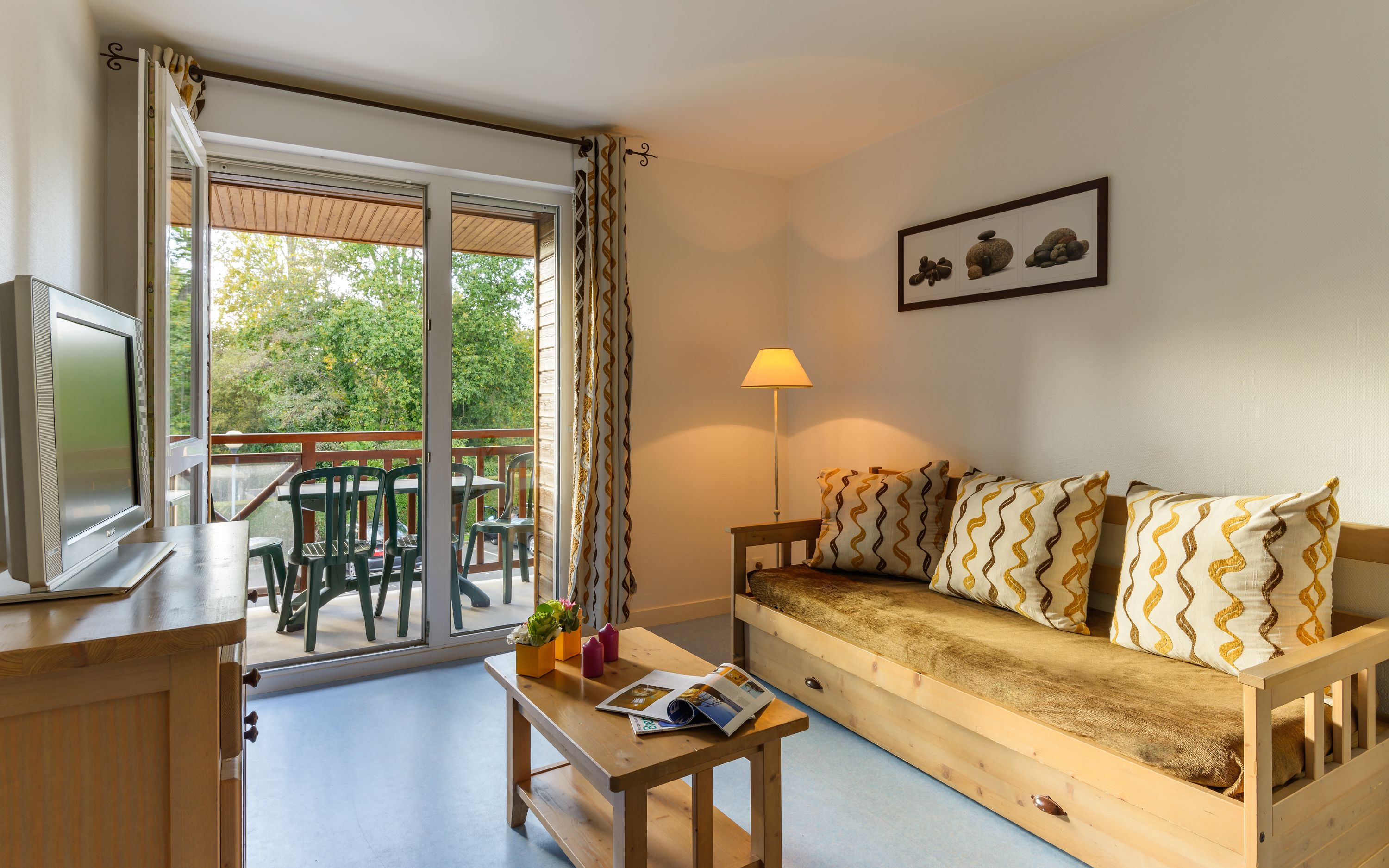 Ein gemütliches Zimmer mit einer Couch, einem Tisch mit einer Zeitschrift, einem Fernseher und einem Balkon mit Stühlen.