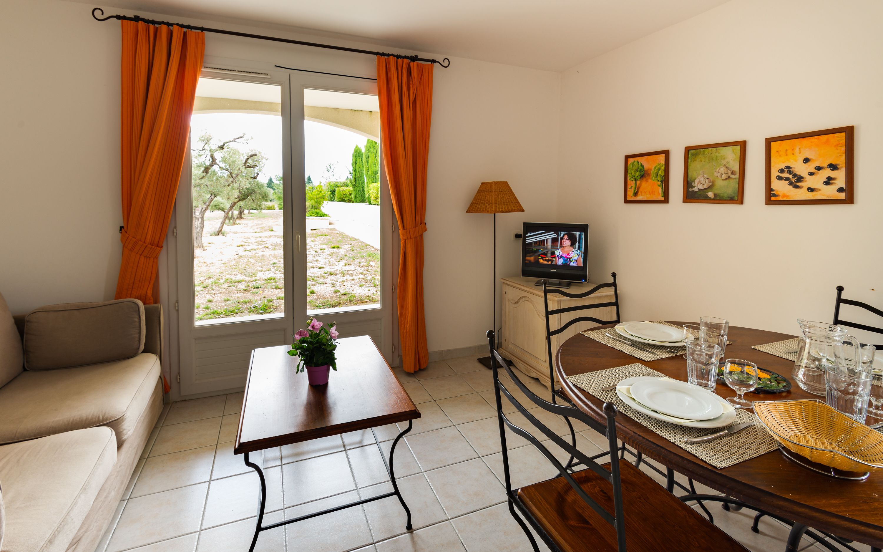 A furnished room with a sofa, a dining table set for two, a TV, and a window with curtains.