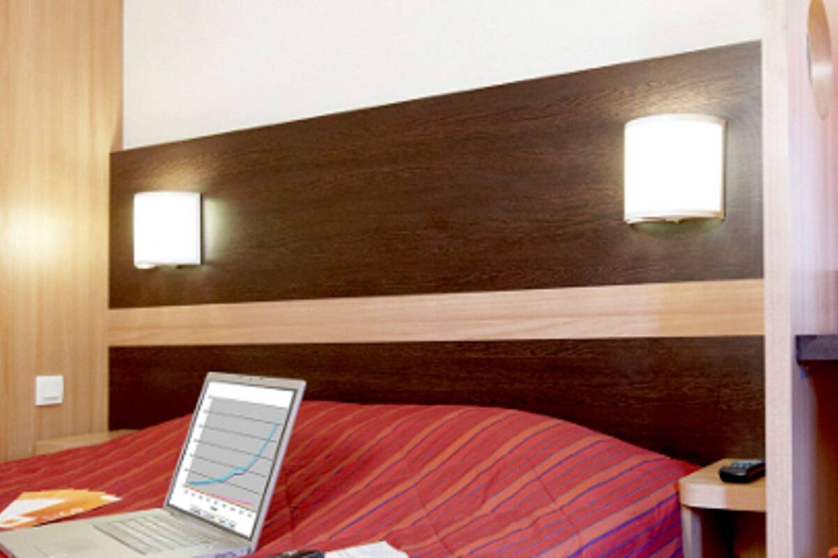 Une chambre d’hôtel avec un lit, un ordinateur portable posé sur le lit, une lampe et une table de chevet.
