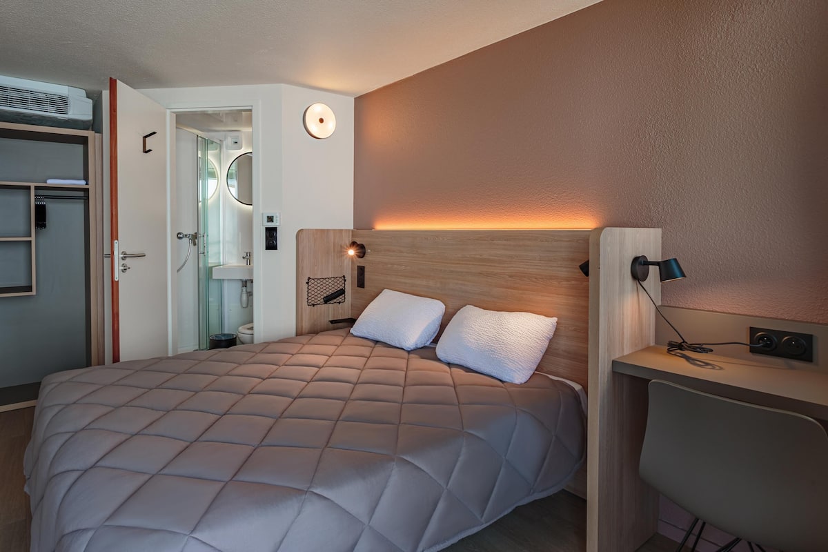 A modern hotel room with a bed, bedside tables, a desk, and a shower area.