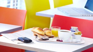 A breakfast tray with bread, juice, and a small container, a mobile phone, and a newspaper on a table with a laptop in the background. - Premiere Classe Montpellier Sud - Lattes (Lattes)