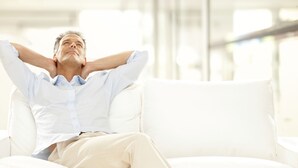 A person sitting on a white couch in a bright room with large windows.