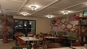 A restaurant with multiple tables and chairs, a mural on the wall, and ceiling lights.