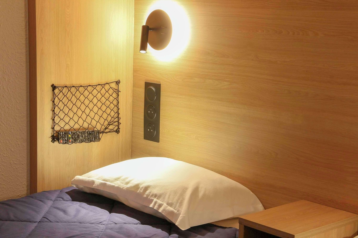 Un dormitorio con cabecera de madera, una cama con almohada blanca, una mesita de noche de madera y una lámpara fijada a la pared.