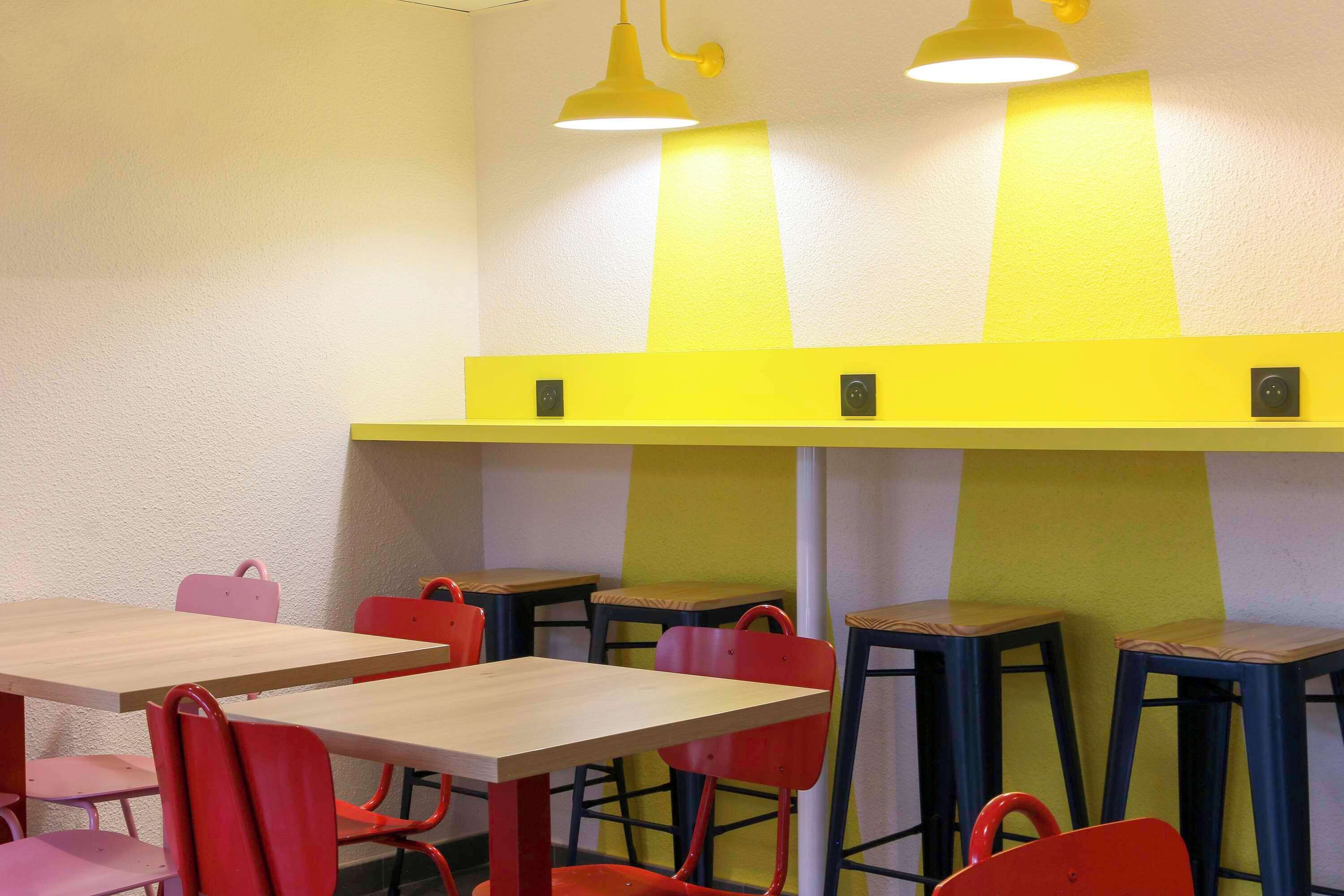 Un comedor con mesas de madera y sillas rojas, paredes de acento amarillo e iluminación colgante.