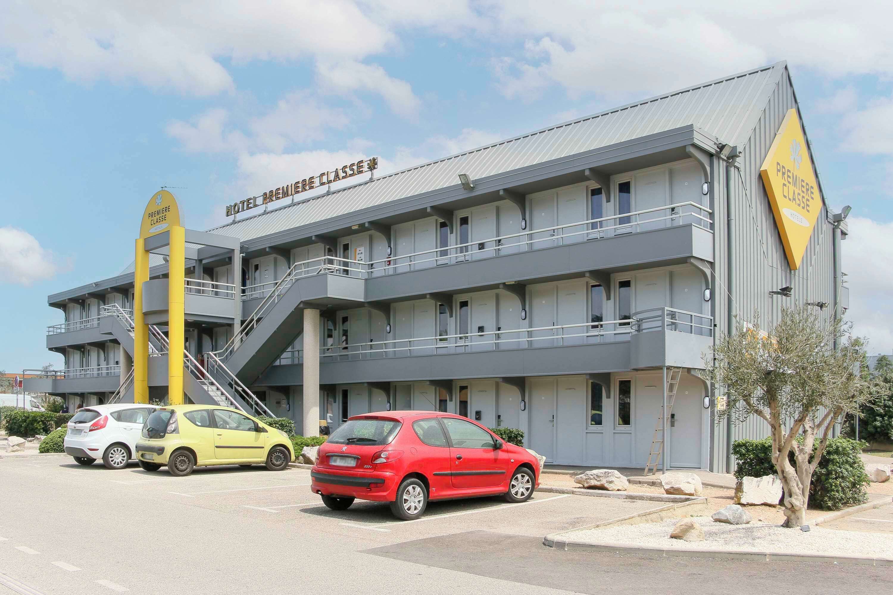 Un hotel con un cartello che riporta la scritta "PREMIERE CLASSE" e una scalinata che conduce all'ingresso. Due automobili sono parcheggiate davanti.
