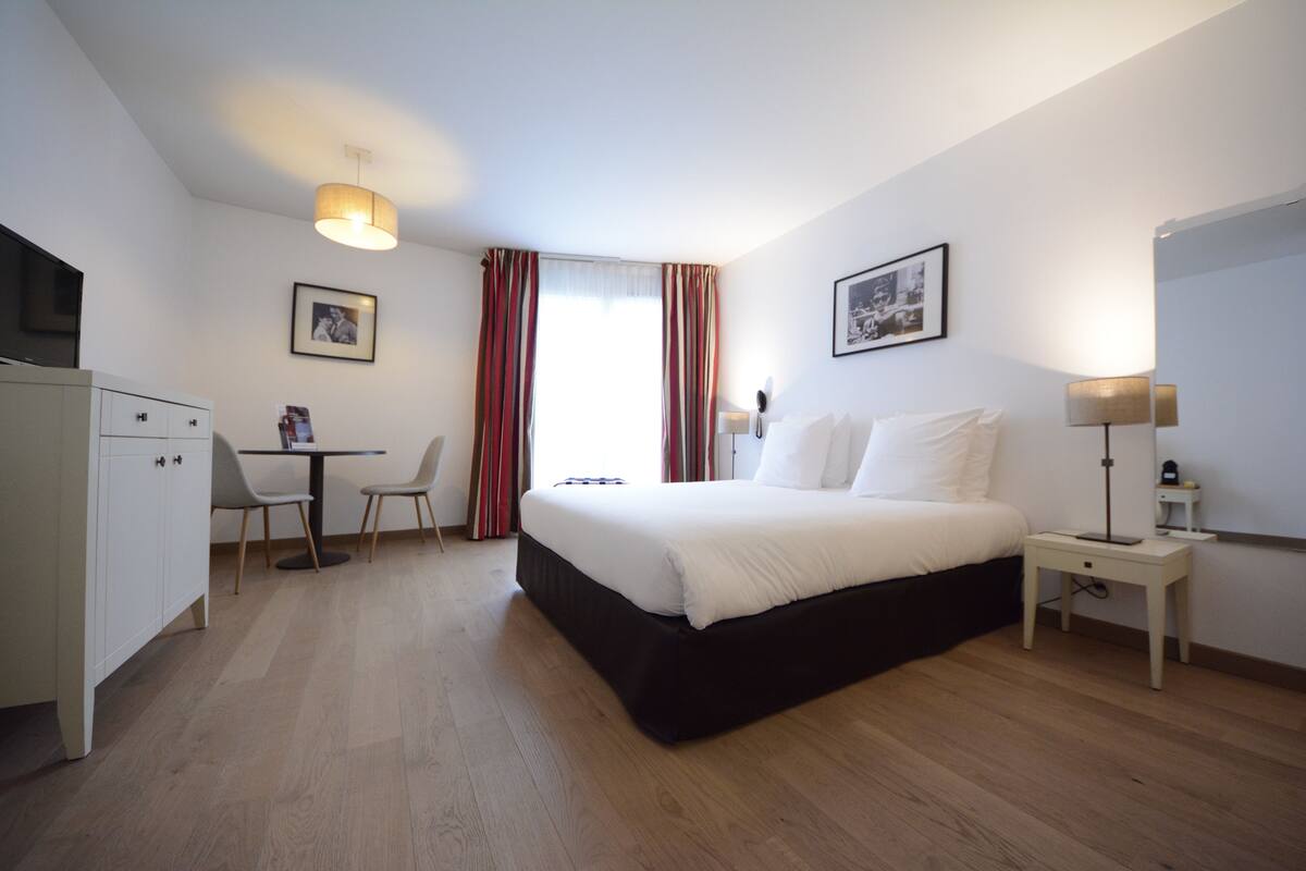 A hotel room with a bed, bedside table, lamp, desk, chairs, and a wardrobe.