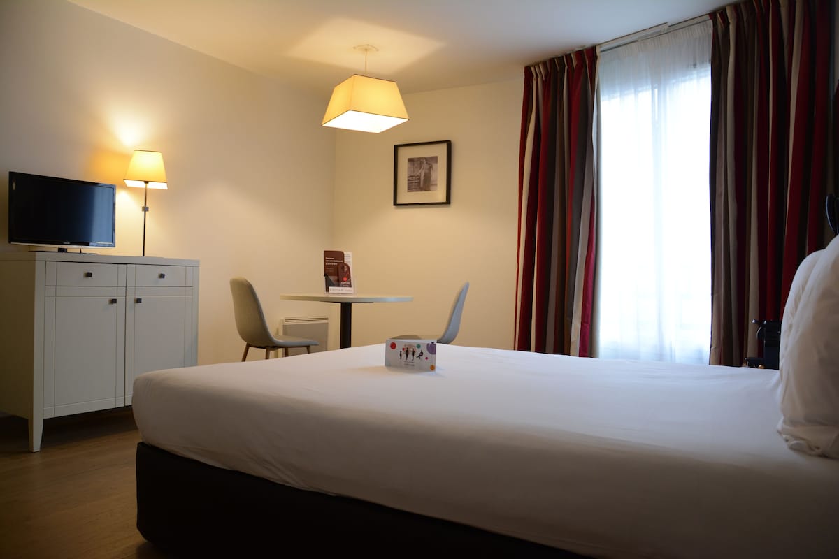 Una habitación de hotel con cama, escritorio, silla, televisión y ventana con cortinas.