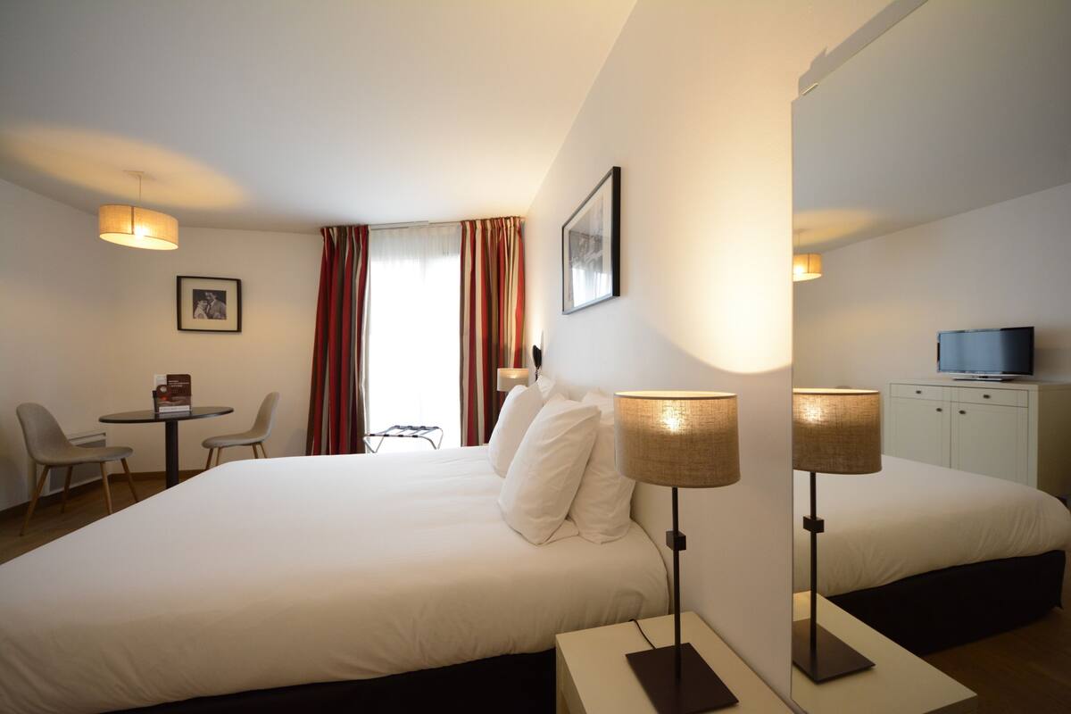 A hotel room with a large bed, two bedside lamps, a desk with chairs, a television, and a framed picture on the wall.