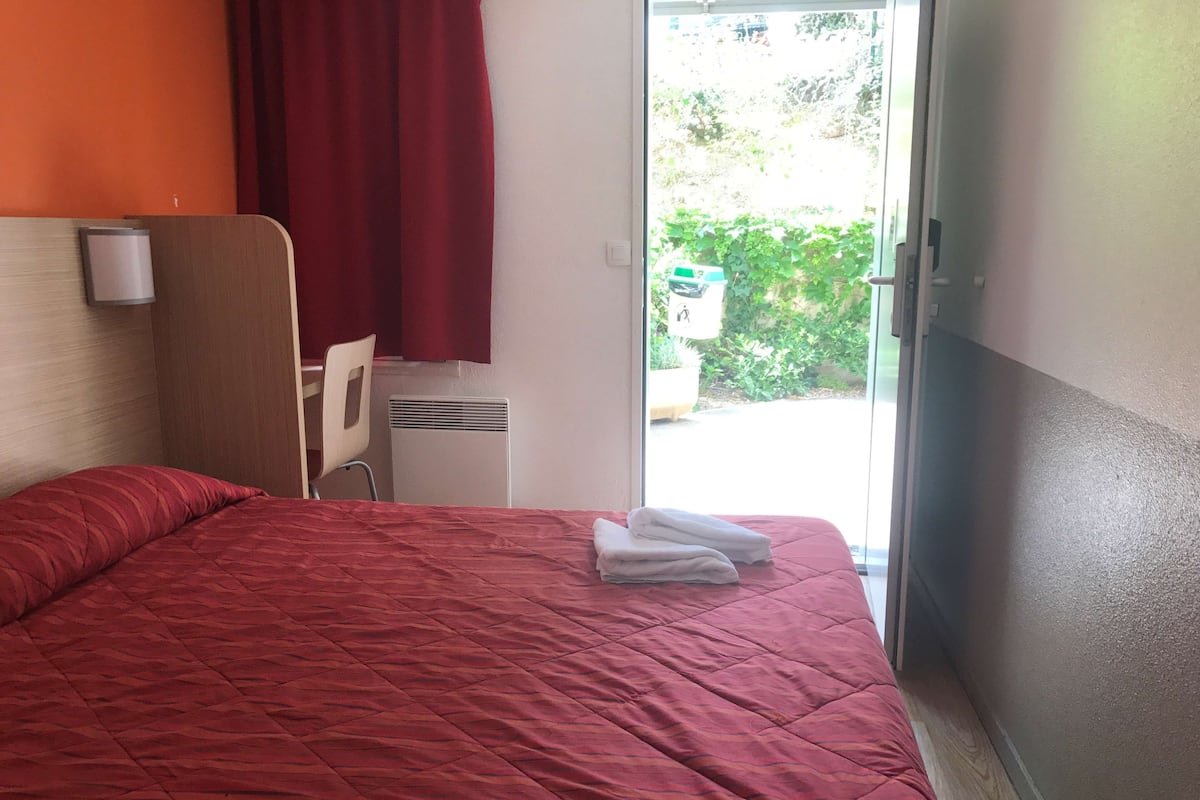 Habitación de hotel con colcha roja, cabecera de madera, una silla y una ventana con vistas al exterior, lleno de vegetación.