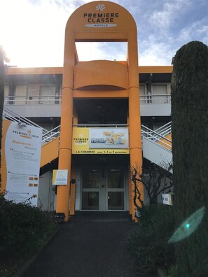 L’entrée d’un hôtel, avec une arche imposante sur laquelle on peut lire l’inscription « HÔTEL PREMIÈRE CLASSE ».