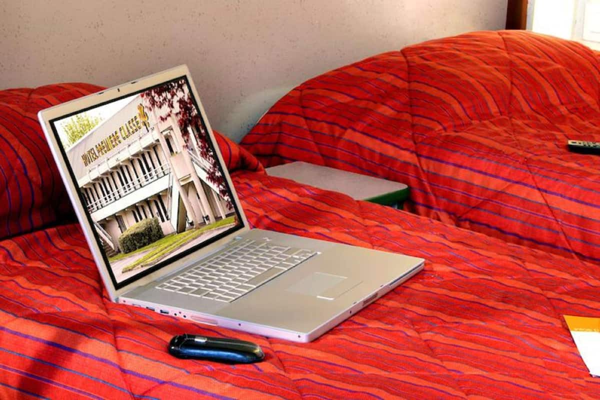 Una laptop sobre una cama con una manta roja, un control remoto y un folleto.