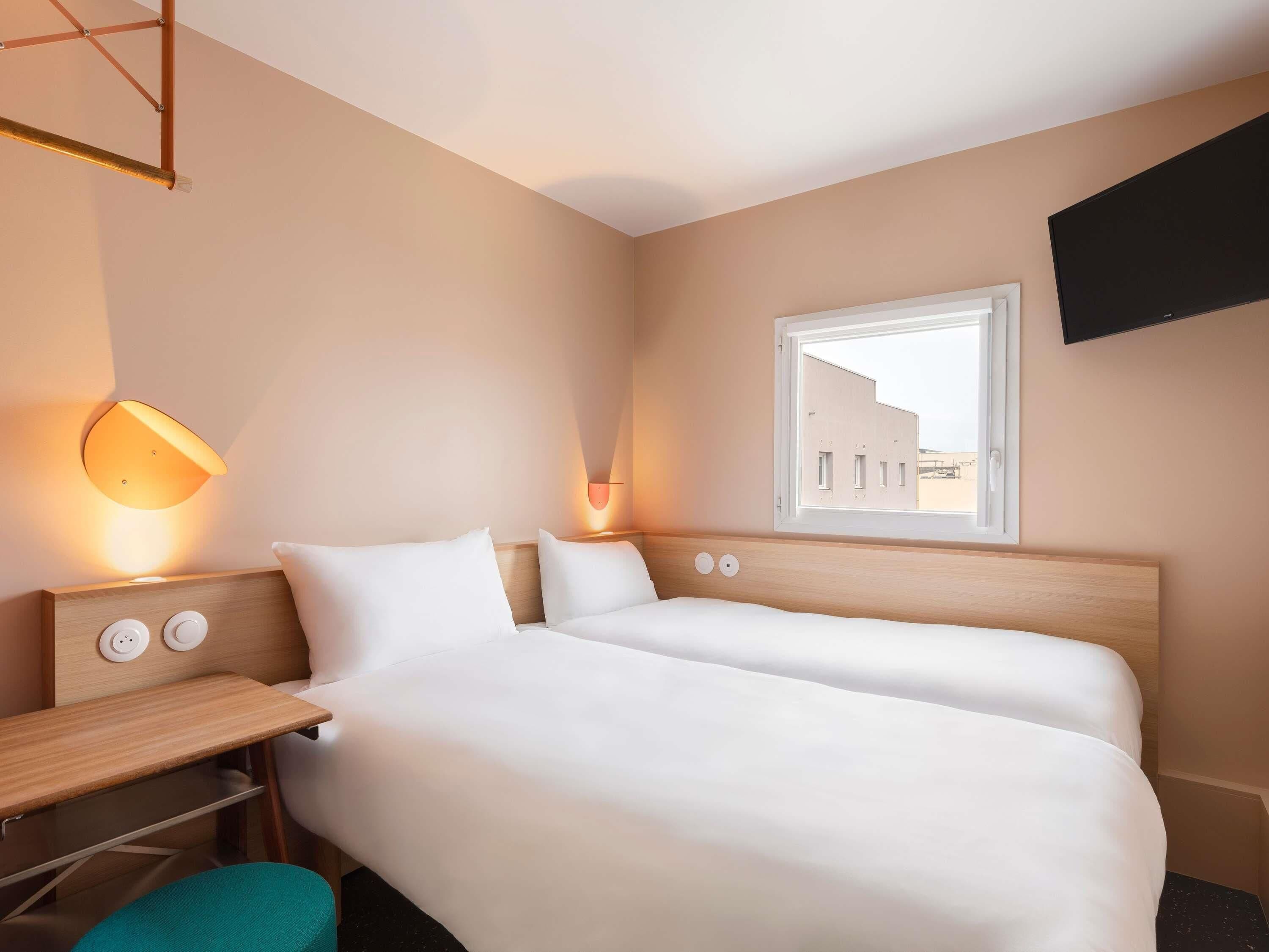 A hotel room with two beds, a desk, a chair, a TV mounted on the wall, and a window with a view of buildings outside.