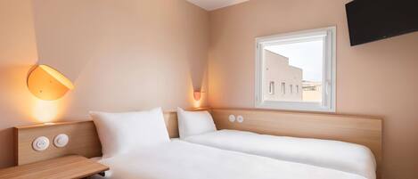A hotel room with two beds, a desk, a chair, a TV mounted on the wall, and a window with a view of buildings outside.