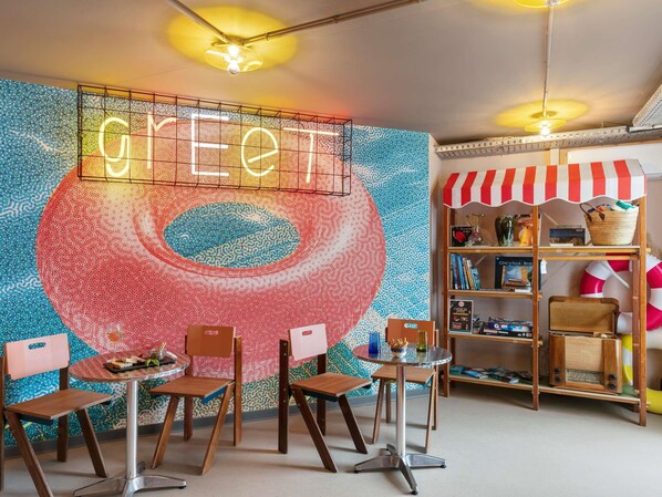 A room with a neon 'GREET' sign, a colorful wall mural, a small round table with chairs, and a bookshelf with various items. - Greet Hotel Nice Aéroport Promenade des Anglais (Nice)