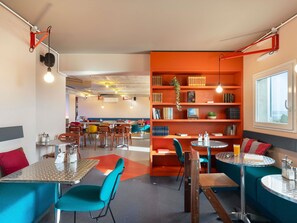 A modern cafe with multiple seating areas, including tables and chairs, and a bookshelf filled with books. - Greet Hotel Nice Aéroport Promenade des Anglais (Nice)