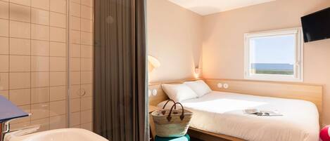 A hotel room with a shower, a sink, a bed with a view of the sea, and a TV mounted on the wall.