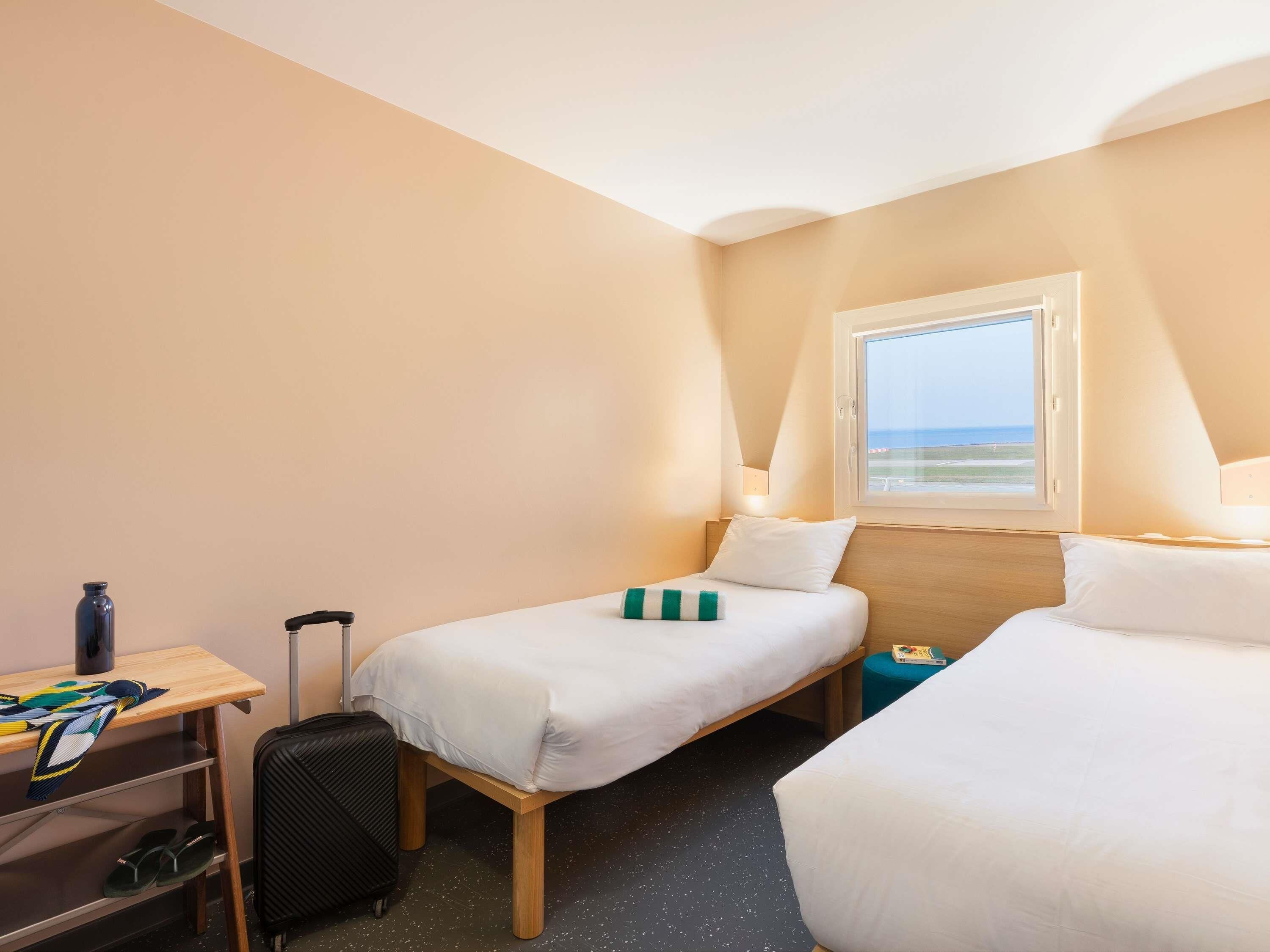 A hotel room with two single beds, a small desk with a chair, a suitcase, and a window with a view of the beach.