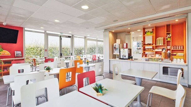 Un restaurante moderno con mesas y sillas blancas, una zona de cocina con armarios color naranja y ventanas amplias que ofrecen vistas a la vegetación exterior.