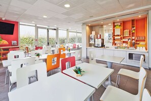 Un restaurant moderne avec des tables et des chaises blanches, un espace cuisine doté de meubles orange et de grandes fenêtres offrant une vue sur la verdure environnante.