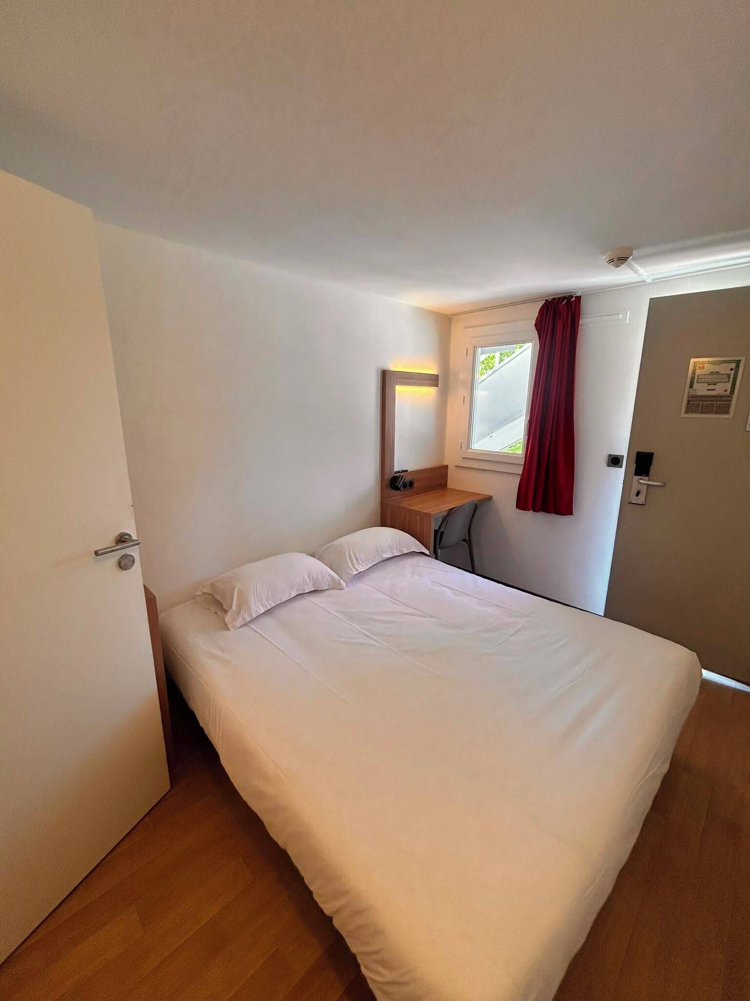 A single bed with white bedding, a wooden nightstand with a mirror, a small desk with a chair, a window with red curtains, and a door with a handle.