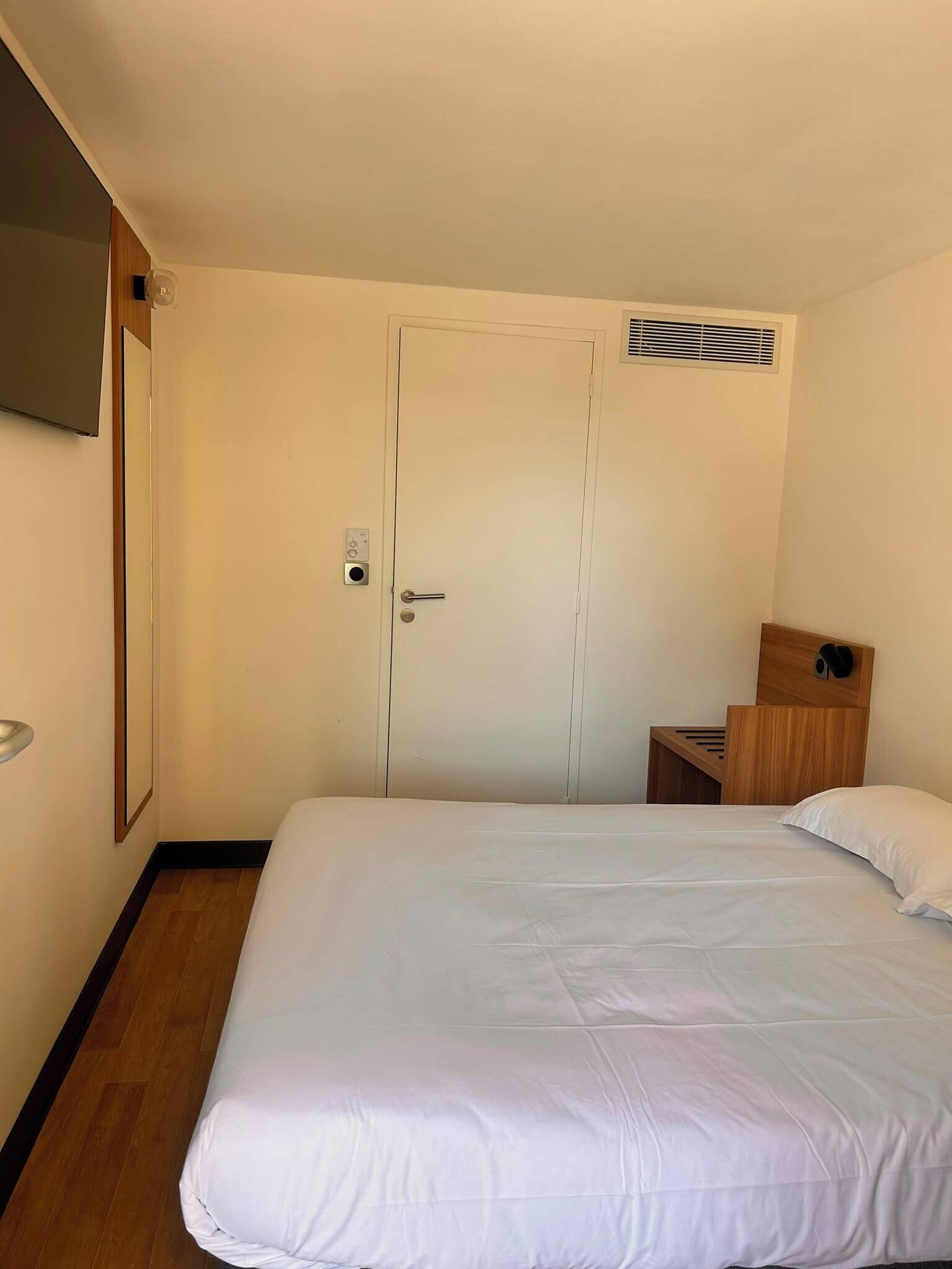 A hotel room with a bed, a door, a small wooden nightstand, a mirror, and a vent.