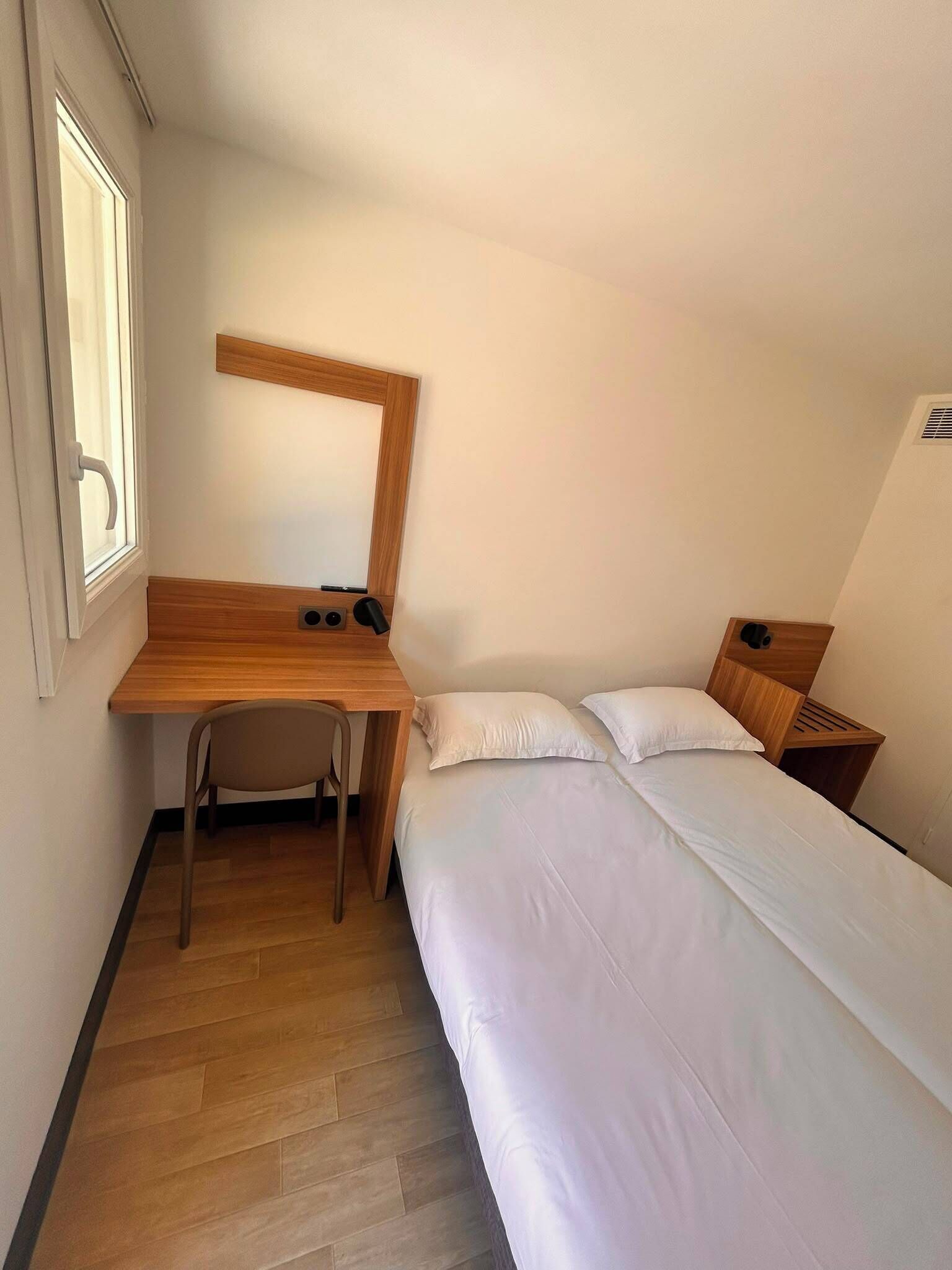 A small, single-room hotel room with a wooden desk, a chair, a bed with white bedding, and a window.