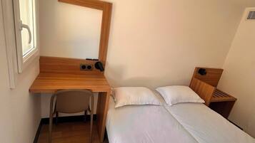 A small, single-room hotel room with a wooden desk, a chair, a bed with white bedding, and a window.