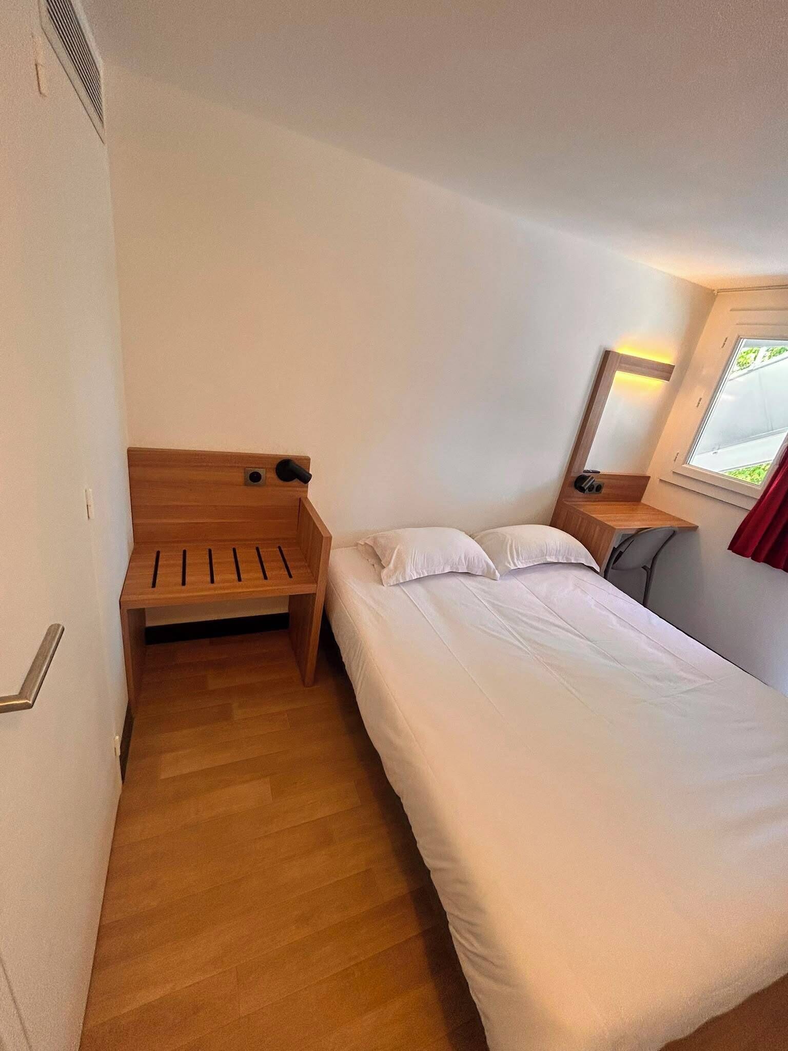 A small, single-bed room with a wooden bench, a bedside table, a mirror, and a window with curtains.