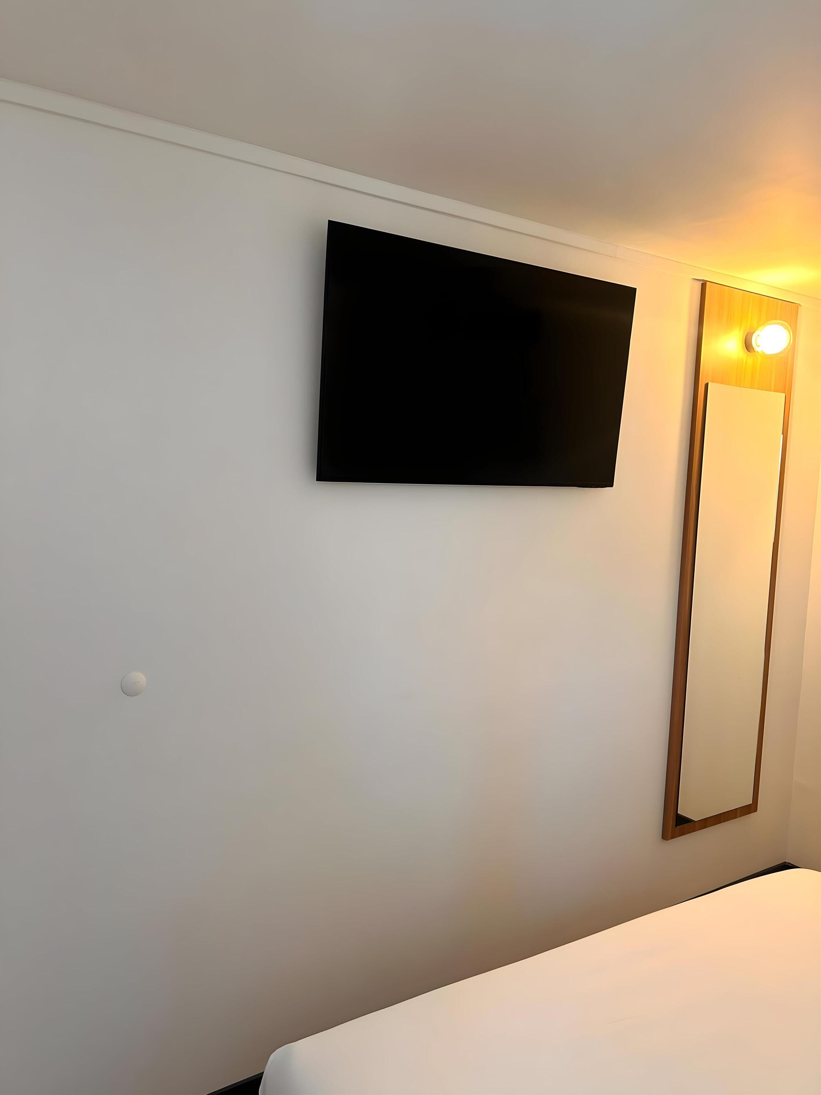 A room with a bed, a wall-mounted TV, and a mirror with a light.