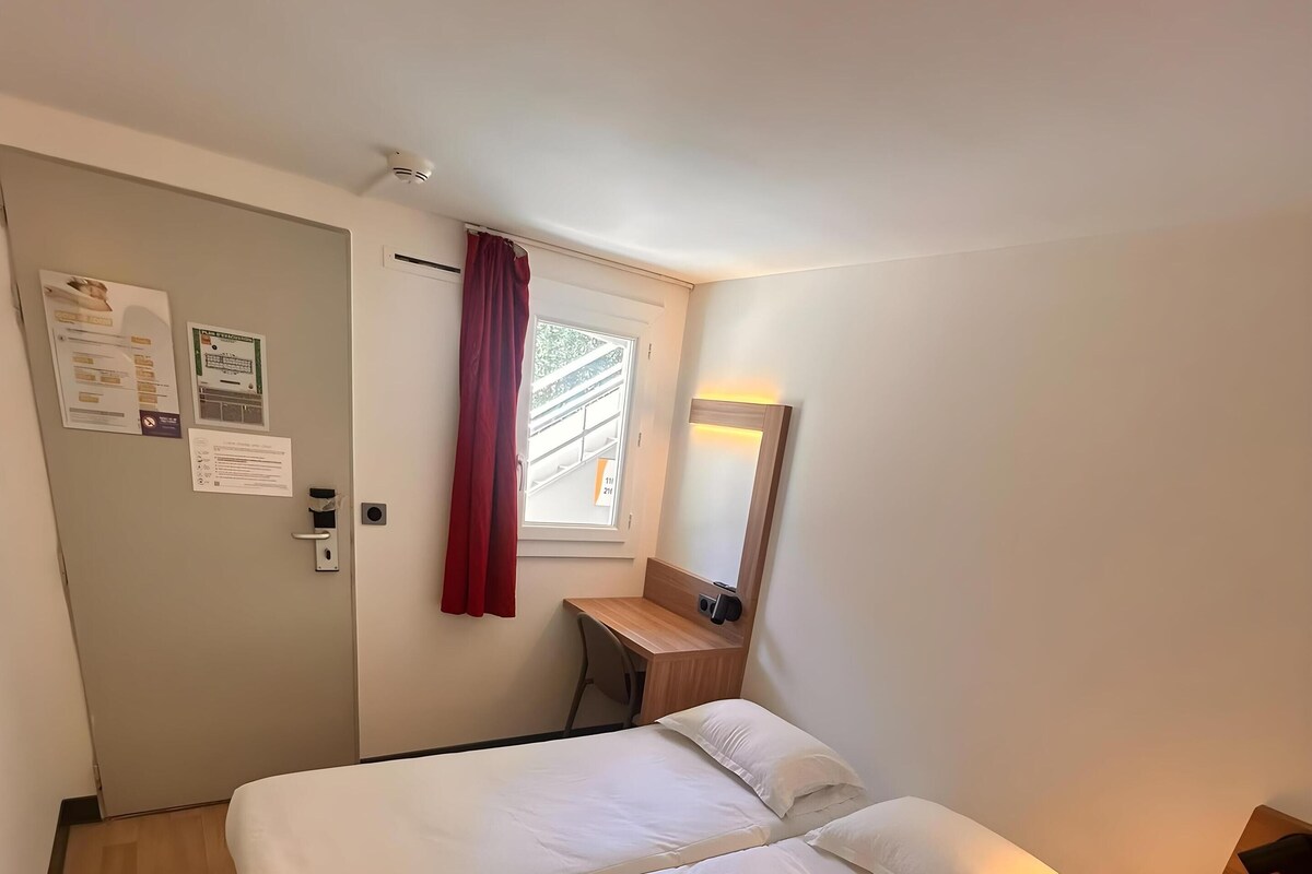 A room with a bed, a desk, a chair, a window with a curtain, and a wall-mounted mirror.