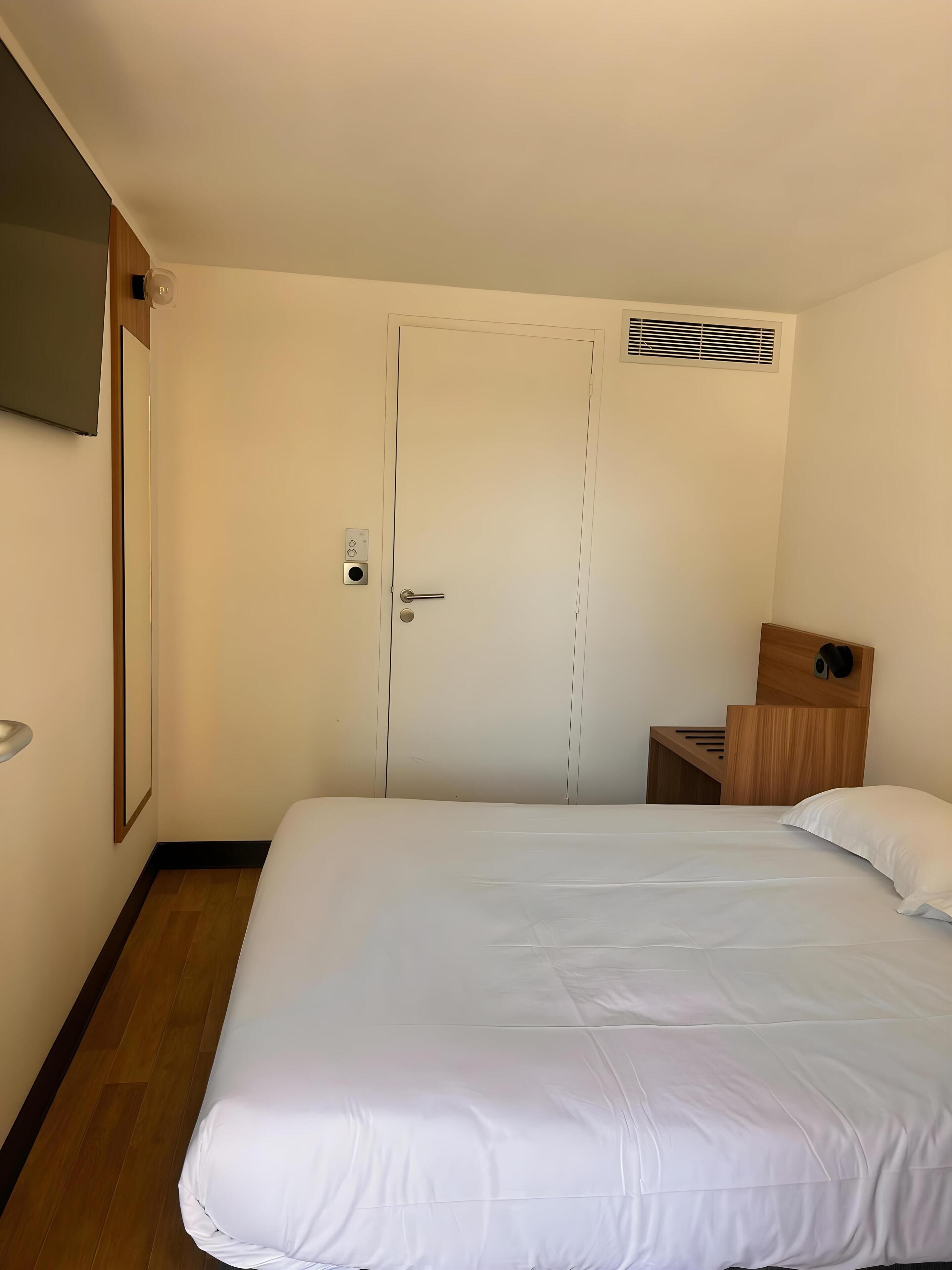 Una habitación de hotel con una cama individual, una puerta con manija, una mesita de noche pequeña y un aplique de pared.