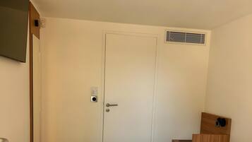 Una habitación de hotel con una cama individual, una puerta con manija, una mesita de noche pequeña y un aplique de pared.